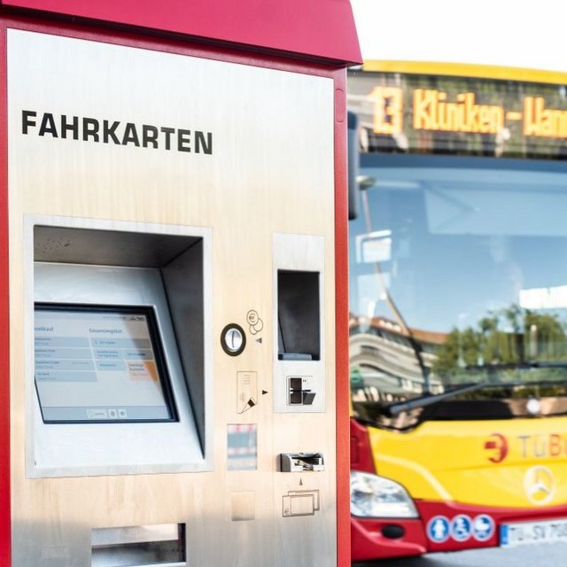 Ein Fahrkartenautomat in Rot und Silber steht an einer Haltestelle. Der Bildschirm zeigt Informationen zur Ticketauswahl an. Auf der linken Seite des Automaten befinden sich verschiedene Bedienelemente, darunter ein Kartenlesegerät und eine Geldautomateneinheit. Im Hintergrund ist ein Bus zu sehen, dessen Haltestellenanzeige die nächste Station anzeigt.