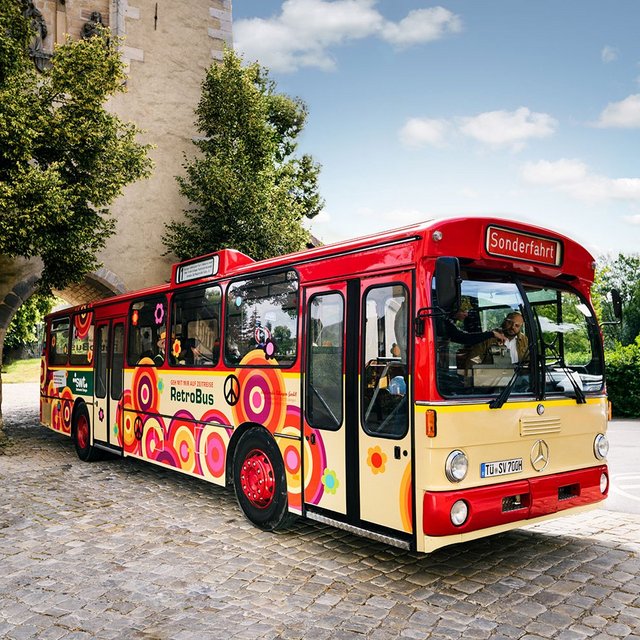 Ein buntes, retro gestyltes Linienbus steht an einer gepflasterten Straße, umgeben von Bäumen und historischer Architektur. Der Bus hat ein rotes Dach und ist mit farbenfrohen Kreisen und Blumen verziert. Auf der Frontscheibe ist das Wort "Sonderfahrt" zu lesen. Im Hintergrund sind grüne Bäume und ein klarer blauer Himmel zu sehen.