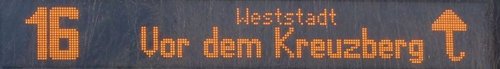 Ein digitaler Anzeigetafel zeigt die Nummer 16 und den Zielort "Vor dem Kreuzberg" an, begleitet von einem Pfeil, der nach oben zeigt. Der Text ist in orange leuchtenden Punkten dargestellt.