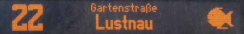 Eine digitale Anzeige mit der Nummer 22, dem Ziel "Gartenstraße" und "Lustnau" in orange leuchtenden Buchstaben, begleitet von einem Symbol, das einen Fisch darstellt.