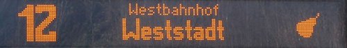Eine digitale Anzeigetafel zeigt die Linie 12, die zum Westbahnhof führt, mit einem orangefarbenen, stilisierten Icon auf der rechten Seite.