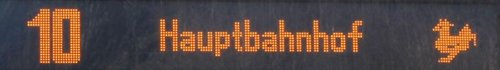 Eine digitale Anzeige zeigt die Zahl "10" und das Wort "Hauptbahnhof" in leuchtenden orangefarbenen Zeichen. Rechts davon befindet sich ein kleines Symbol, das einen Huhn darstellt.