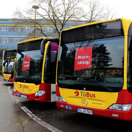 Drei Linienbusse mit der Aufschrift "TüBus" stehen geparkt. Auf jedem Bus sind rote Schilder mit den Worten "Warnstreik!" und "Heute!" sichtbar. Im Hintergrund ist ein modernes Gebäude mit Glasfassade zu sehen, und die Bäume sind noch kahl, was auf Frühling hindeutet. Die Umgebung ist städtisch, mit einer Fahrbahn und einer Parkfläche.