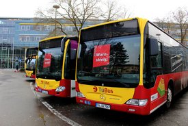 Drei Linienbusse mit der Aufschrift "TüBus" stehen geparkt. Auf jedem Bus sind rote Schilder mit den Worten "Warnstreik!" und "Heute!" sichtbar. Im Hintergrund ist ein modernes Gebäude mit Glasfassade zu sehen, und die Bäume sind noch kahl, was auf Frühling hindeutet. Die Umgebung ist städtisch, mit einer Fahrbahn und einer Parkfläche.