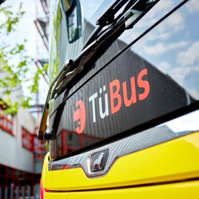 Ein gelber Bus mit dem Schriftzug "TüBus" auf der schwarzen Rückseite ist in der Nähe eines Gebäudes abgebildet. Im Hintergrund sind grüne Bäume und eine Struktur zu sehen, die hervorgehoben wird von hellem Licht. Die Perspektive ist leicht schräg, was den Vordergrund des Busses betont.