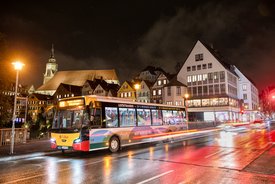 Ein Stadtbus steht an einer Haltestelle in einer Stadt bei Nacht. Im Hintergrund sind mehrstöckige Gebäude mit verschiedenen Fassaden zu sehen, darunter Fachwerkhäuser. Ein Kirchturm ragt über die Dächer hinaus. Die Straßen sind nass von Regen, reflektieren das Licht der Straßenlaternen und der Fahrzeuglichter. Der Bus hat eine bunte Lackierung und zeigt die Liniennummer 198.
