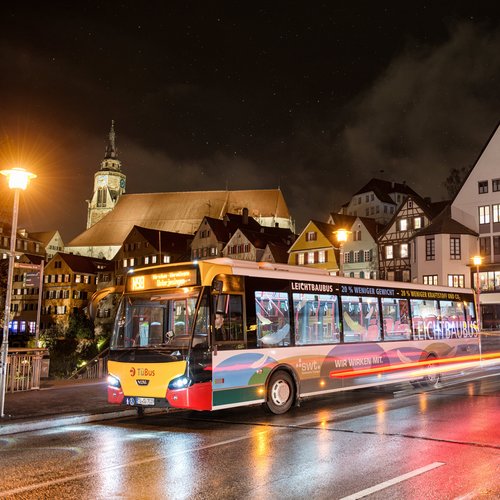 Ein Stadtbus steht an einer Haltestelle in einer Stadt bei Nacht. Im Hintergrund sind mehrstöckige Gebäude mit verschiedenen Fassaden zu sehen, darunter Fachwerkhäuser. Ein Kirchturm ragt über die Dächer hinaus. Die Straßen sind nass von Regen, reflektieren das Licht der Straßenlaternen und der Fahrzeuglichter. Der Bus hat eine bunte Lackierung und zeigt die Liniennummer 198.