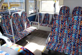 Das Bild zeigt das Innere eines Busses mit bunten, gemusterten Sitzen. Die Sitzpolster sind in lebhaften Farben wie Rot, Blau, Gelb und Schwarz gestaltet, mit einem geometrischen Muster aus kleinen Quadraten. Der Boden des Busses ist aus dunklem Holzmaterial, und es gibt metallische Haltegriffe an den Sitzen. Im Hintergrund sind Fenster, durch die Tageslicht hereinströmt, sowie eine Ansicht von draußen mit parkenden Autos und Geschäften.