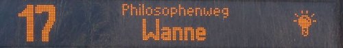 Ein elektronisches Anzeigegerät zeigt die Nummer 17 und die Namen "Philosophenweg" und "Wanne" in leuchtenden orangen Zeichen an. Rechts ist ein Symbol, das eine Lichtquelle darstellt. Der Hintergrund ist dunkel.
