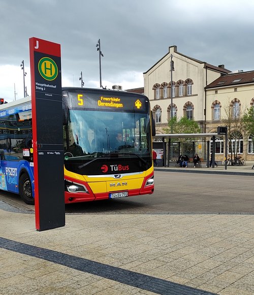 Eine Straßenansicht mit einem roten und gelben Bus von TuBus, der an einer Haltestelle hält. Der Bus hat das Ziel "Feuerbgäde Derendingen" und die Liniennummer 5 angezeigt. Im Hintergrund ist ein historisches Gebäude mit großen Fenstern und einem braunen Dach zu sehen. Es sind einige Bäume und Sitzmöglichkeiten in der Umgebung, sowie ein weiterer Bus in blauer Farbe, der an der Haltestelle steht. Der Himmel ist bewölkt und es herrscht eine ruhige Atmosphäre.