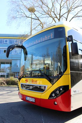 Ein gelb-roter Linienbus steht an einer Haltestelle. Auf der Anzeige des Busses steht "Betriebsfahrt". Der Bus hat eine moderne Form und die Aufschrift "TüBus" auf der Seite. Im Hintergrund sind hohe Gebäude und ein blauer Himmel zu sehen, während Bäume ohne Blätter im Vordergrund stehen.