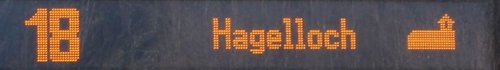 Eine digitale Anzeige mit einer orangefarbenen Schrift. Auf der Anzeige steht die Nummer 18 und der Ort "Hagelloch", gefolgt von einem Piktogramm eines Gebäudes.