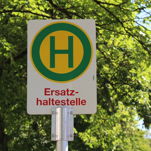 Ein Straßenschild mit einem großen, grünen „H“ in einem gelben Kreis, das auf eine Ersatzhaltestelle hinweist. Der Schriftzug „Ersatzhaltestelle“ ist in roter Schrift direkt unter dem Symbol. Der Hintergrund zeigt hellgrüne Blätter von Bäumen, die im oberen Teil des Bildes sichtbar sind. Der Himmel ist klar und blau.