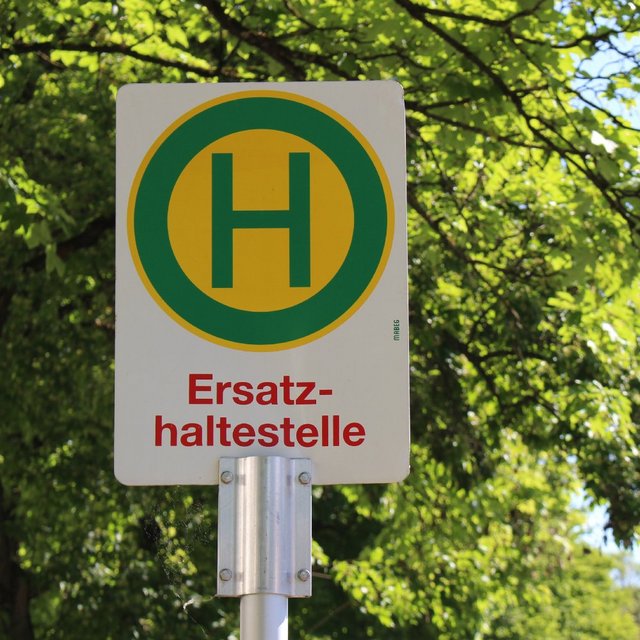 Ein Straßenschild mit einem großen, grünen „H“ in einem gelben Kreis, das auf eine Ersatzhaltestelle hinweist. Der Schriftzug „Ersatzhaltestelle“ ist in roter Schrift direkt unter dem Symbol. Der Hintergrund zeigt hellgrüne Blätter von Bäumen, die im oberen Teil des Bildes sichtbar sind. Der Himmel ist klar und blau.