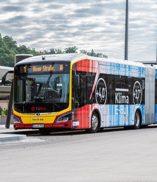 Ein moderner, rot und blau lackierter Linienbus steht an einer Haltestelle. Die Anzeige auf dem Bus zeigt die Route "1" zur "Alxer Straße". Der Bus ist ein E-Bus mit klimafreundlichem Design, das Temperaturen von +4°C und +27°C sowie den Slogan "Wir können nicht bleiben" zeigt. Im Hintergrund sind einige Gebäude und eine andere Fahrzeug auf einer weitläufigen, gepflasterten Fläche zu erkennen. Der Himmel ist bewölkt, und die Umgebung ist von Bäumen umgeben.